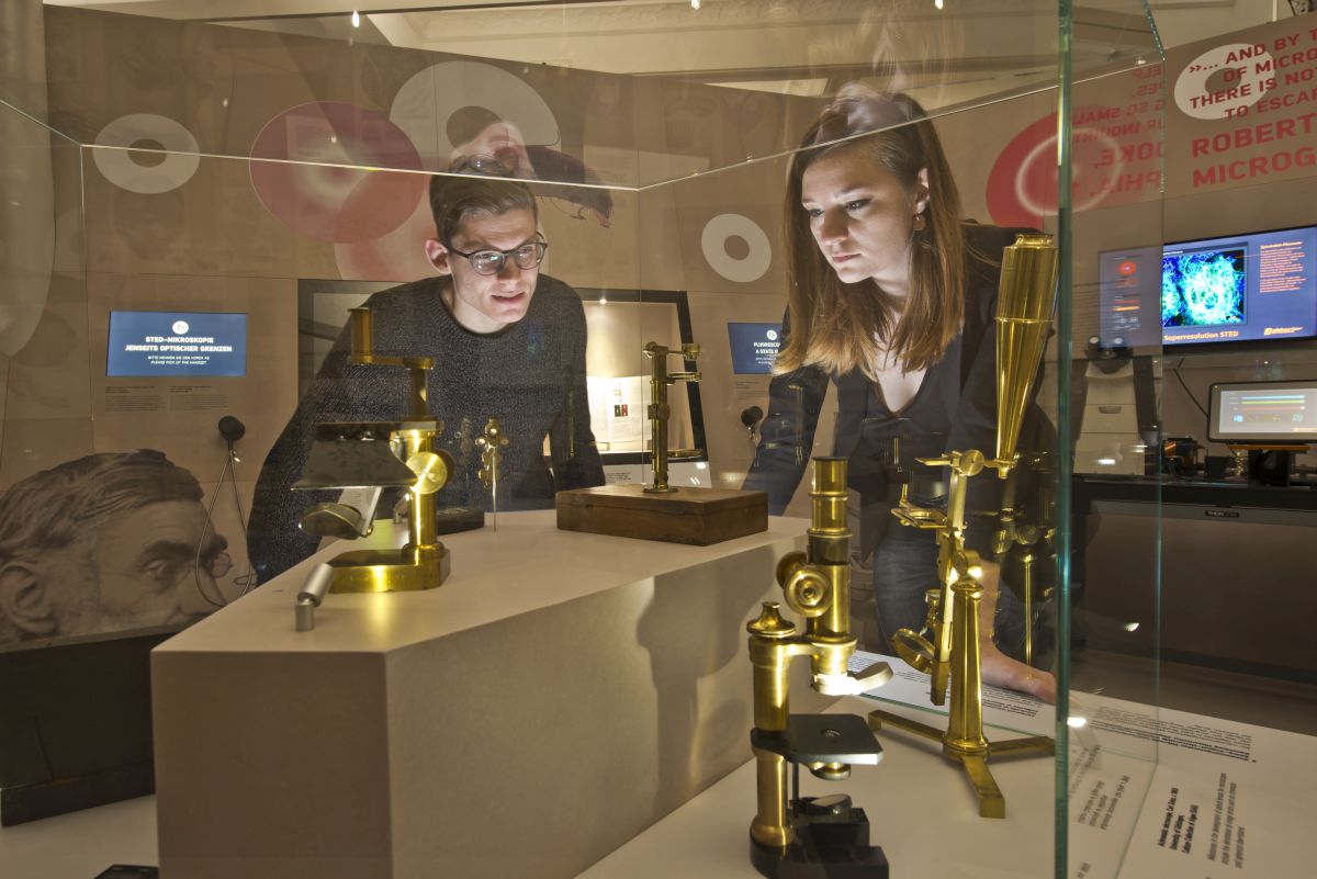 MUseumsbesucher betrachten Mikroskope in einer Glasvitrine.