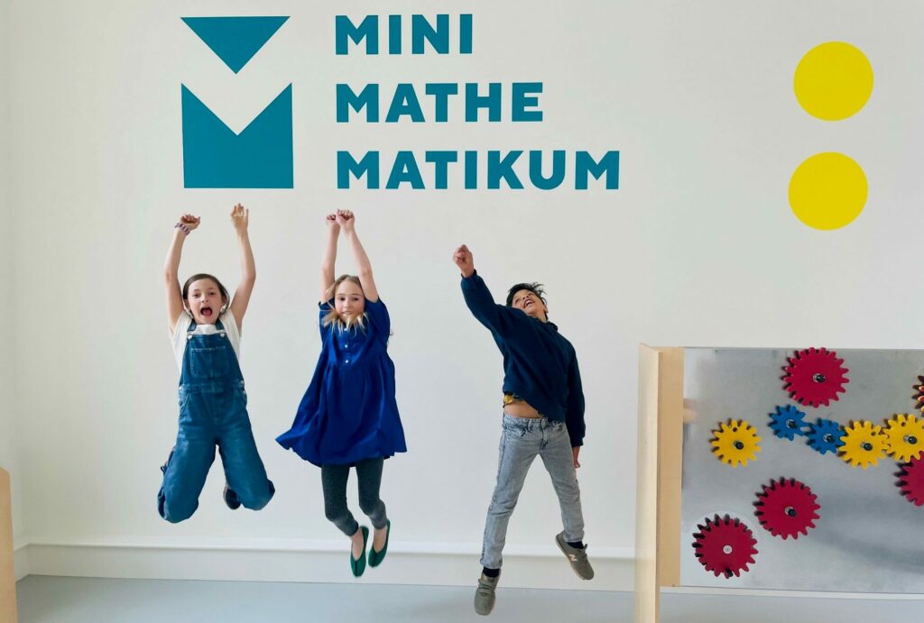 Drei Kinder springen vor einer Wand in die Luft, auf der Mini-Mathematikum steht.
