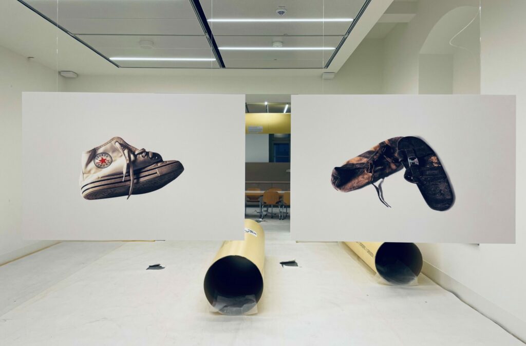 Vorschau-Bild zur Ausstellung: Zwei Bilder, auf denen verschiedene Schuhe abgebildet sind hängen in einem Raum. Auf dem Boden liegen große Metallrohre.