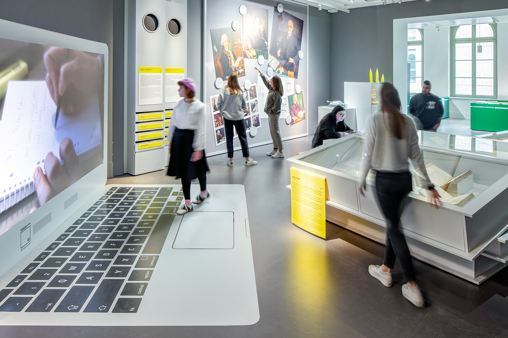 Mehrere Museumsbesucher*innen betrachten Schaukästen und Wand-Installationen im Raum „Schreibtisch“. Im Vordergrund steht das Abbild eines Überlebensgroßen Laptops, dessen Tastatur auf den Boden gemalt ist.