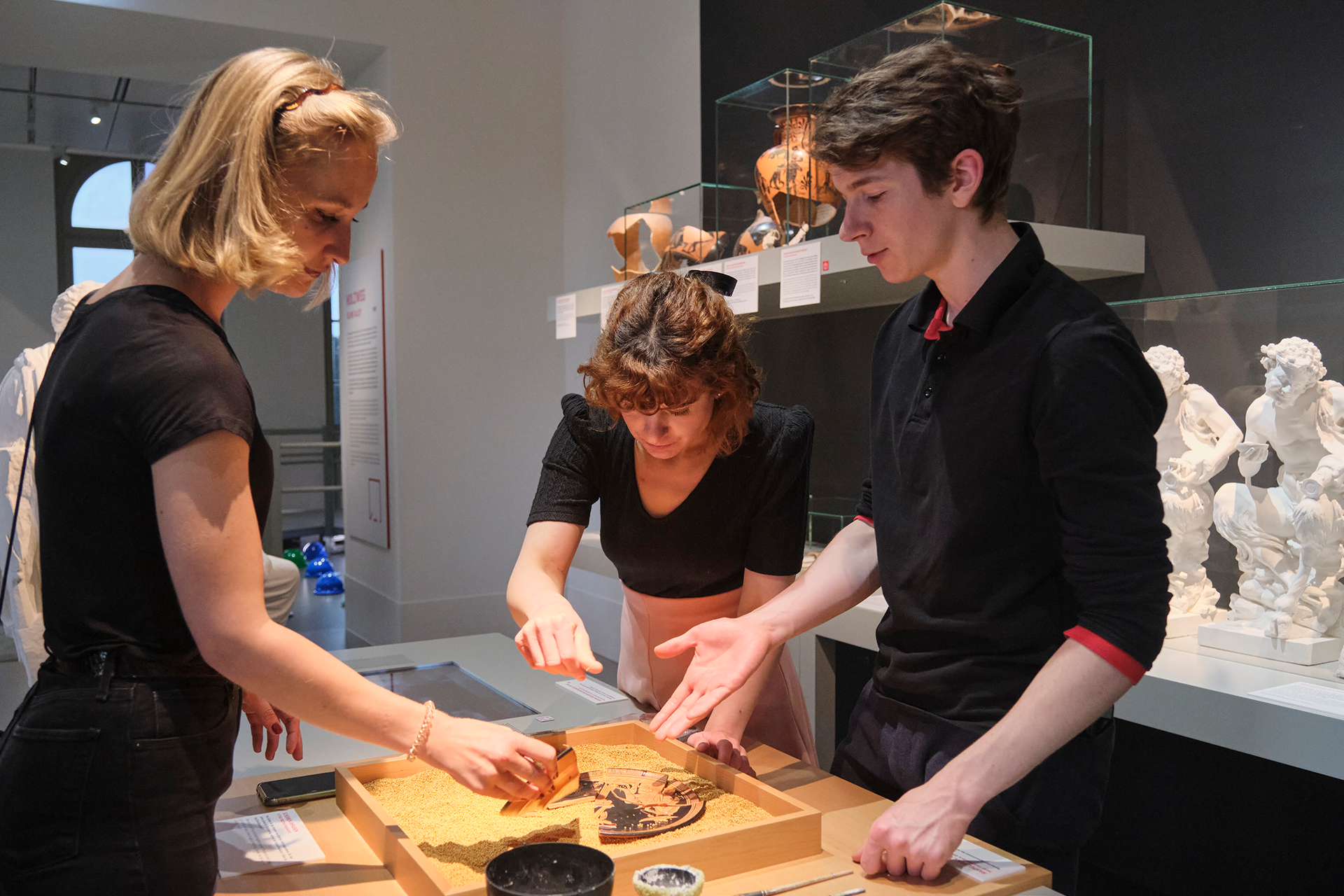 Drei Museumsbesucher*innen stehen an einem Tisch im Raum „Werkstatt“ und versuchen ein Objekt aus Tonscherben zusammenzusetzen.