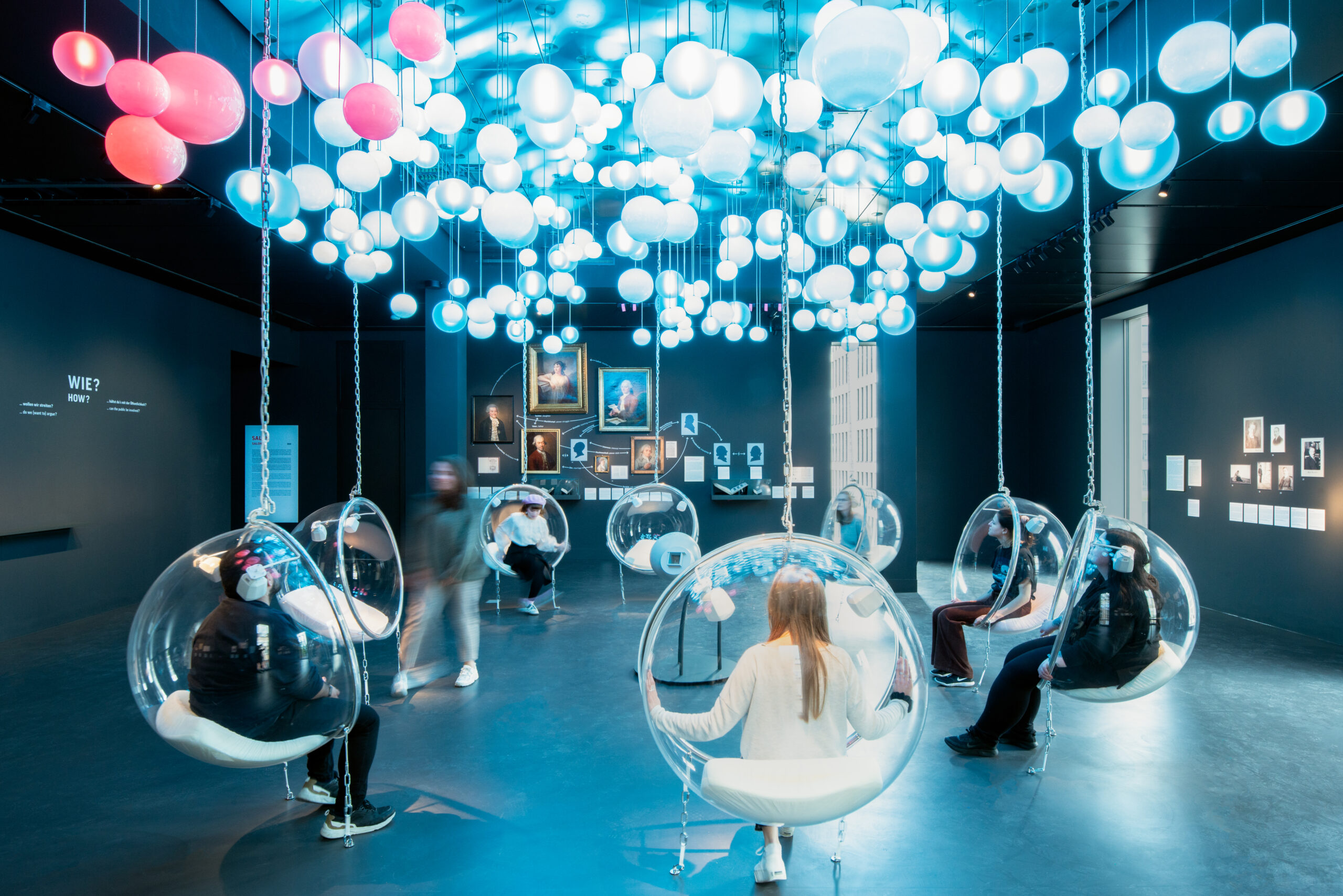 Der Raum „Salon“ ist durch zahlreiche Kugel-Lampen an der Decke beleuchtet. Museumsbesucher*innen sitzen in durchsichtigen, kugelförmigen Hängesesseln und tragen große Kopfhörer. 
