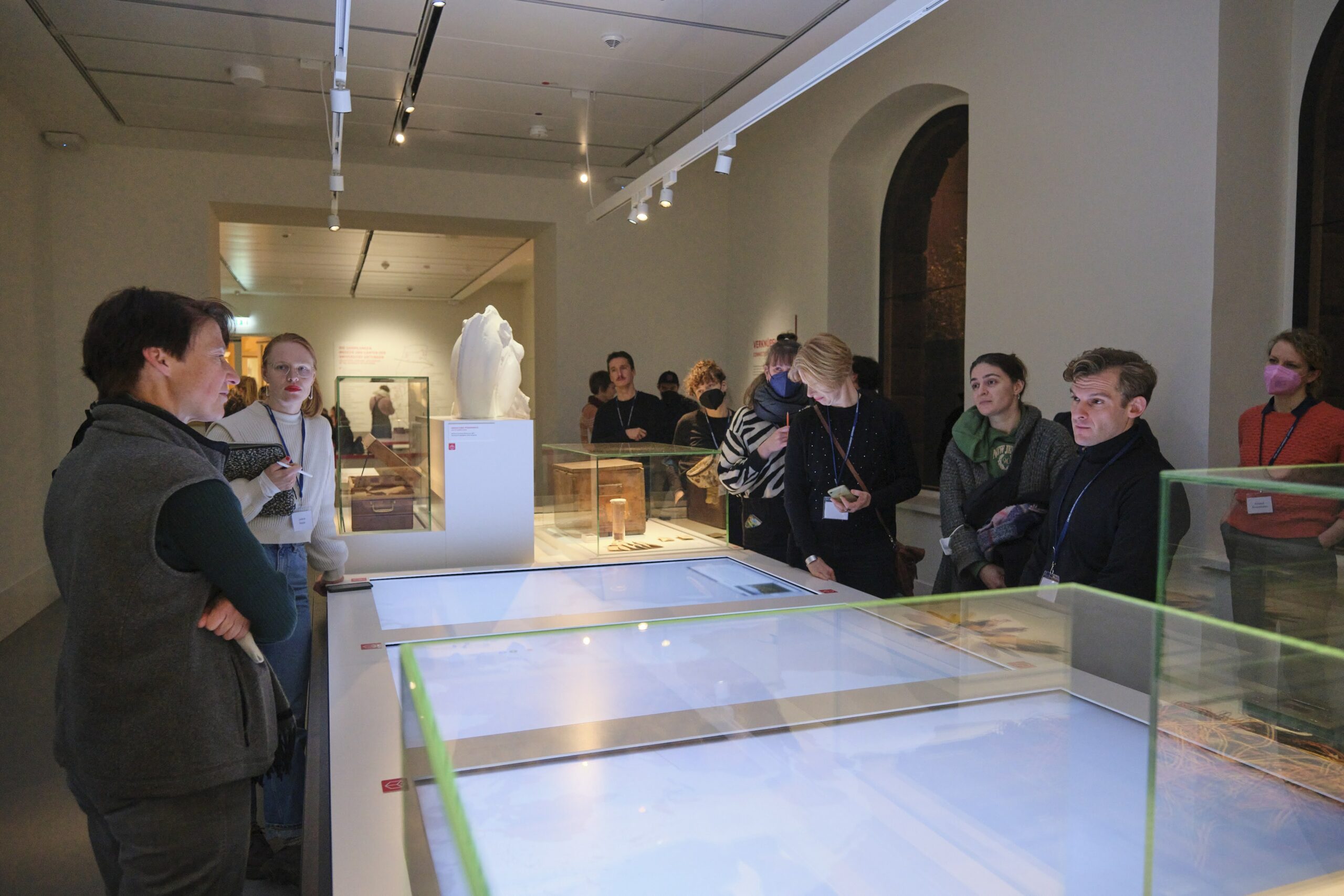Eine Gruppe Museumsbesucher*innen steht um große Monitor-Tische herum.