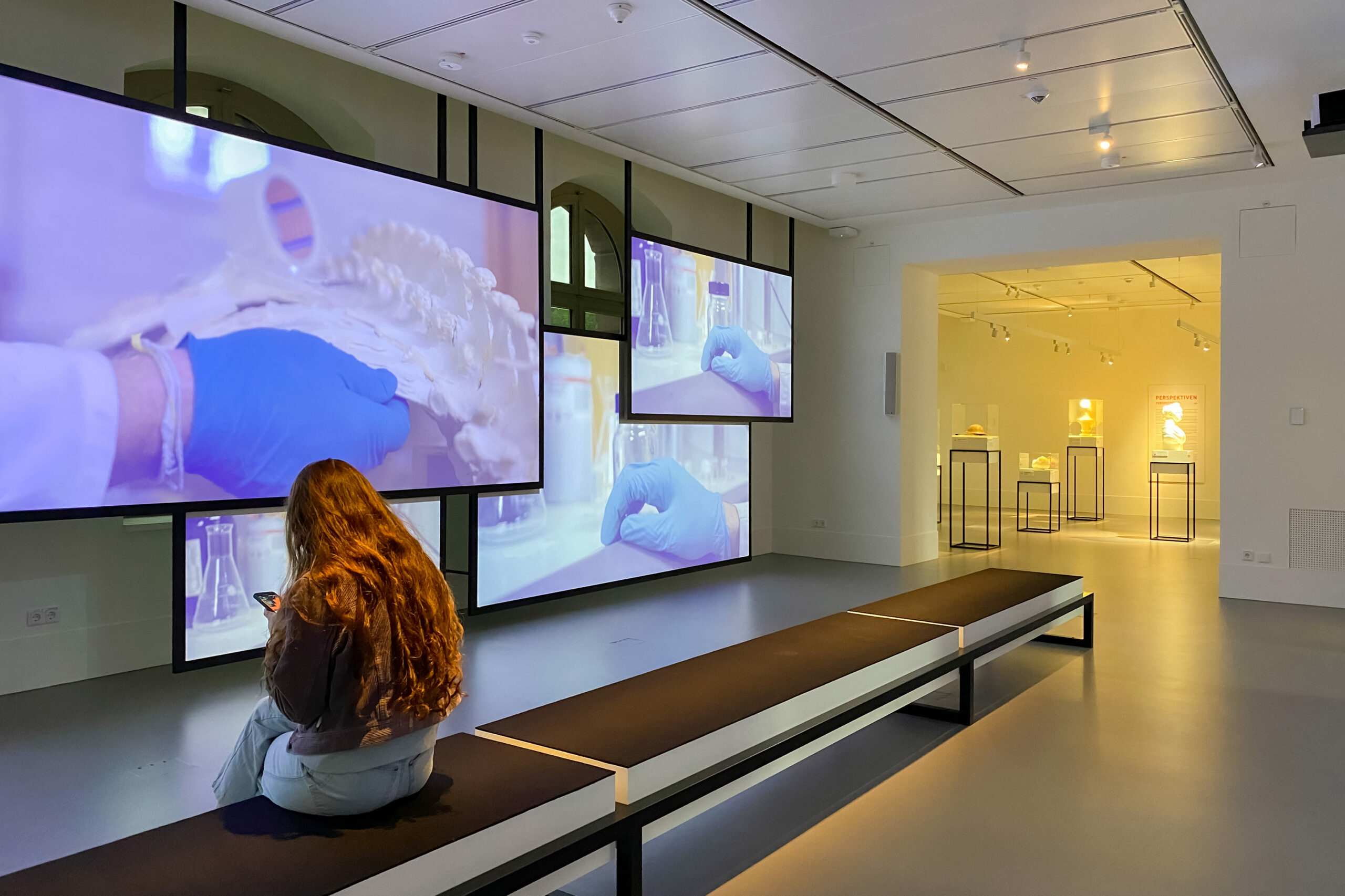 Eine Museumsbesucherin sitzt auf einer Bank vor einer Videoinstallation aus mehreren Bildschirmen.