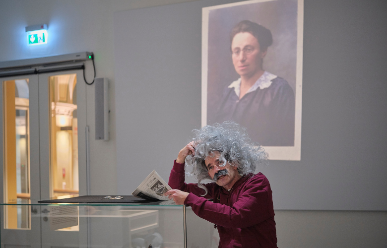 Götz Lautenbach als Albert Einstein mit Perücke und Bart. An eine Vitrine lehnend hält er eine Zeitung in der Hand. Im Hintergrund ist ein Bild von Emmy Noether an die Wand projiziert