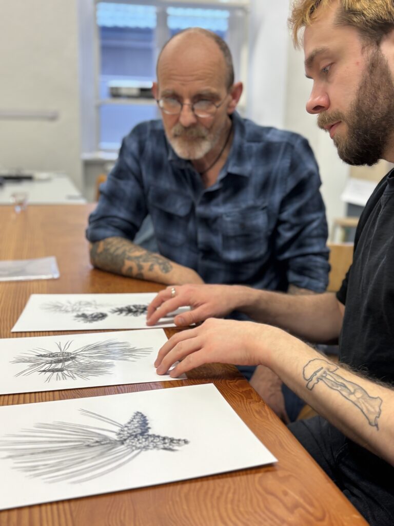 Schwerdtfeger und Gallenkamp sitzen an einem Tisch, vor ihnen liegen die handgezeichneten Tattoovorlagen verschiedener botanischer Motive